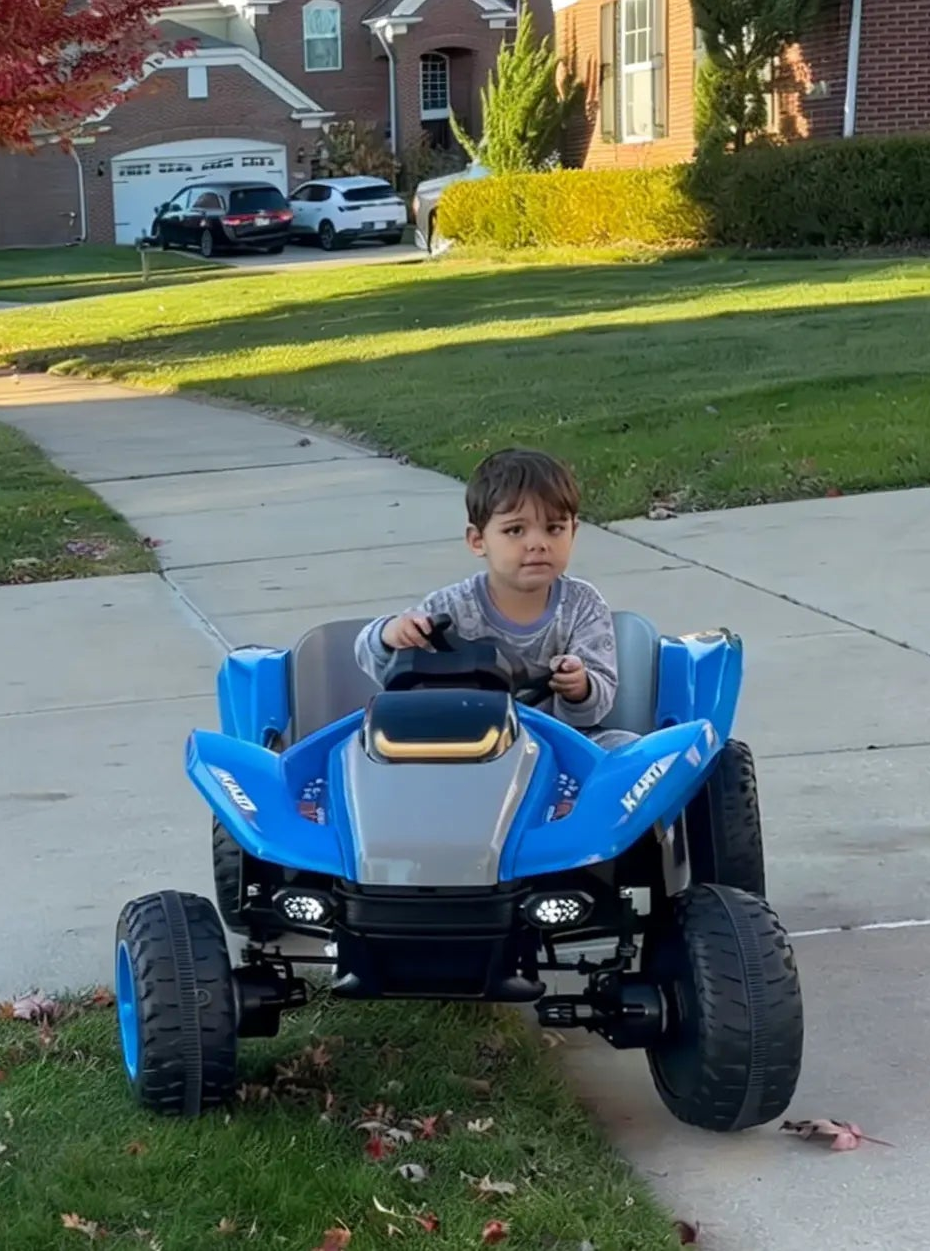 Child sitting in a blue toy car on a residential street with autumn leaves and houses in the background.